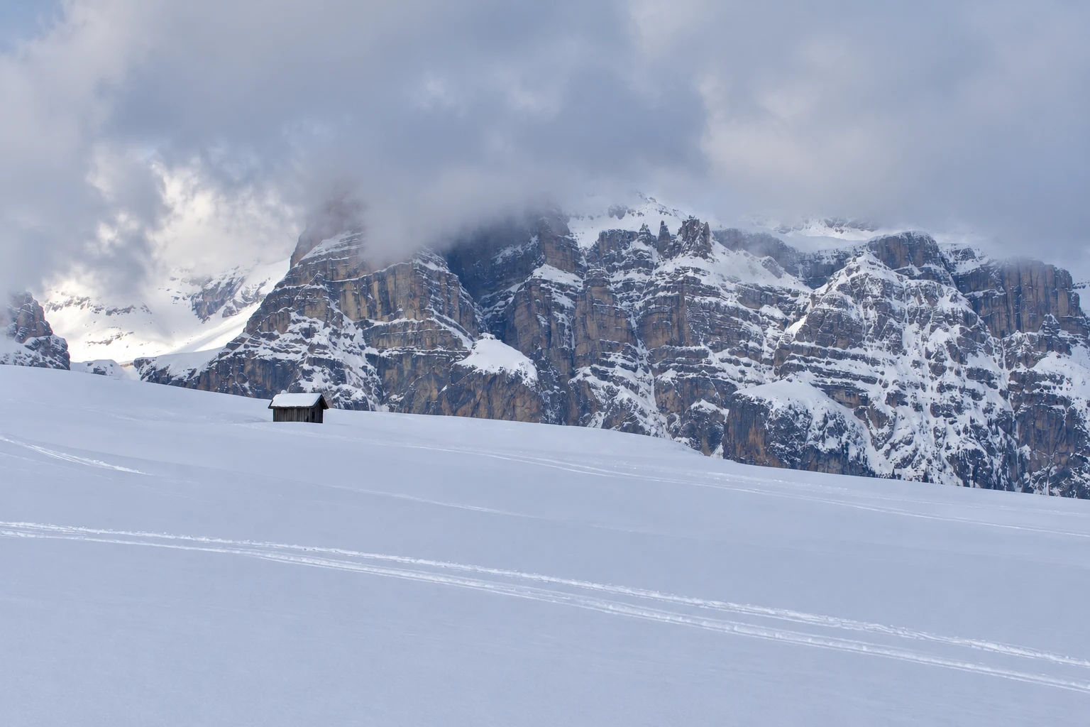 A snowy Dolomite slope below layered limestone cliffs and cloud, with a small timber hut