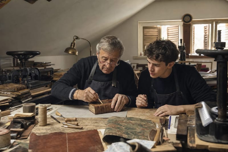 An elderly bookbinder and a young apprentice rebinding an antique leather-bound book at a workbench in a Florence workshop