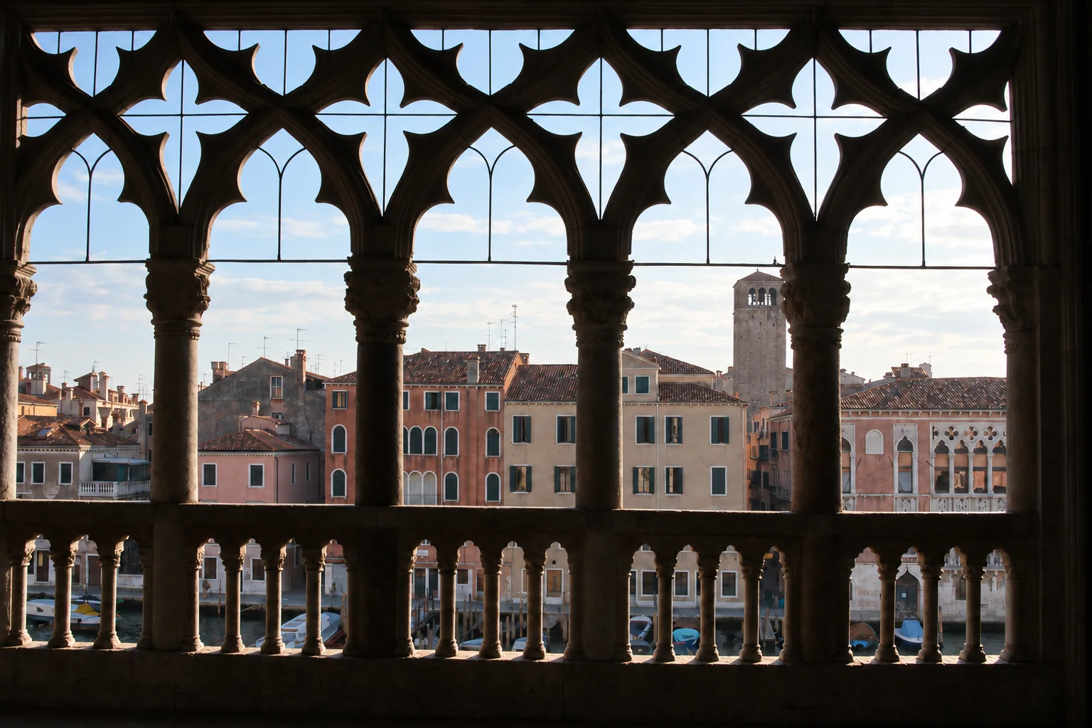 Shadowed Gothic loggia with repeated pointed openings, thin metal bars and a low balustrade framing canal-side rooftops and a tower.