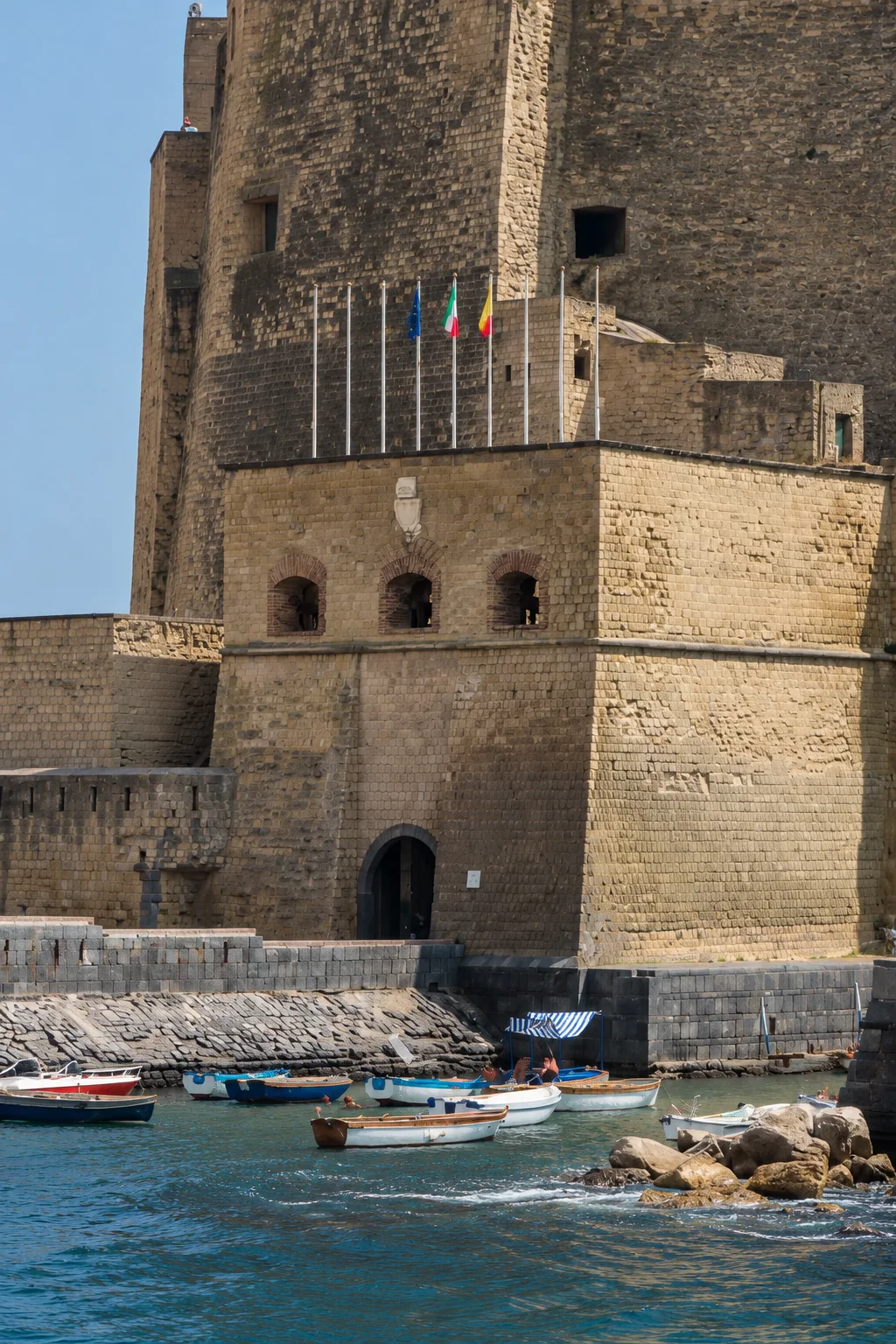 Portrait view of Castel dell’Ovo’s projecting entrance block, with three upper arches, terrace flagpoles, a dark lower doorway and stone seawall.