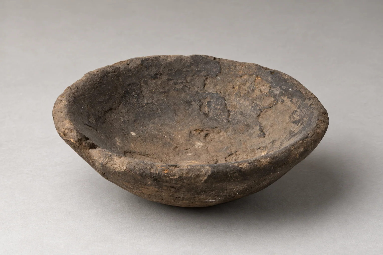 Shallow gray-brown clay bowl with an uneven chipped rim, visible interior cracks and muted ochre patches.