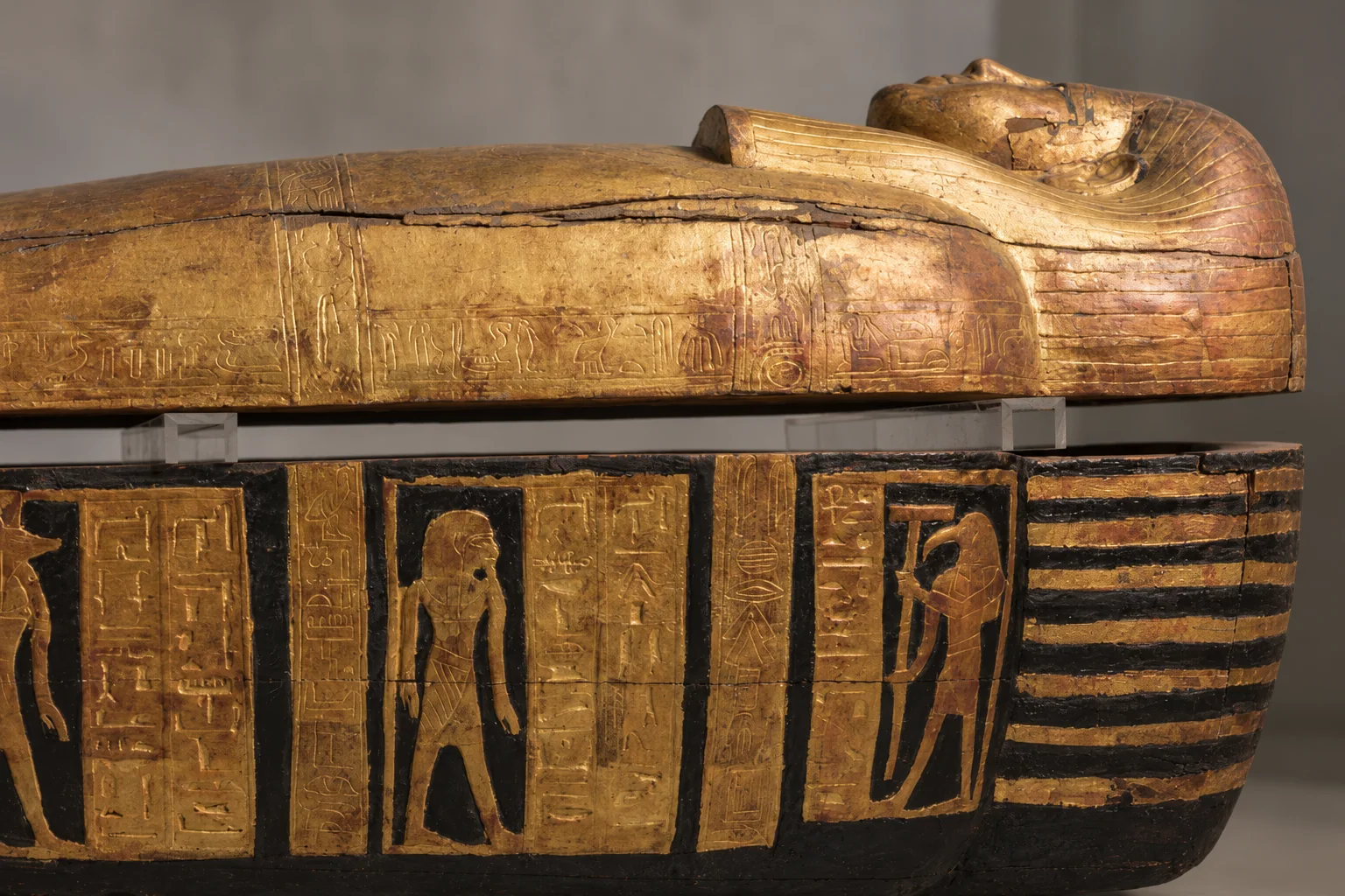 Side view of a worn gilded anthropoid coffin lid, with an upturned profile at the right, above a dark trough with gold figures.
