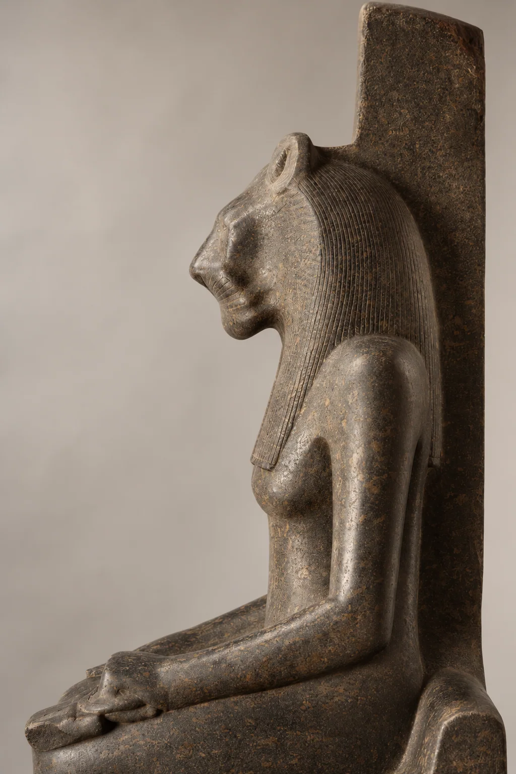 Left-facing lioness head, striated wig, torso and bent arm resting on the lap of a dark speckled stone statue.