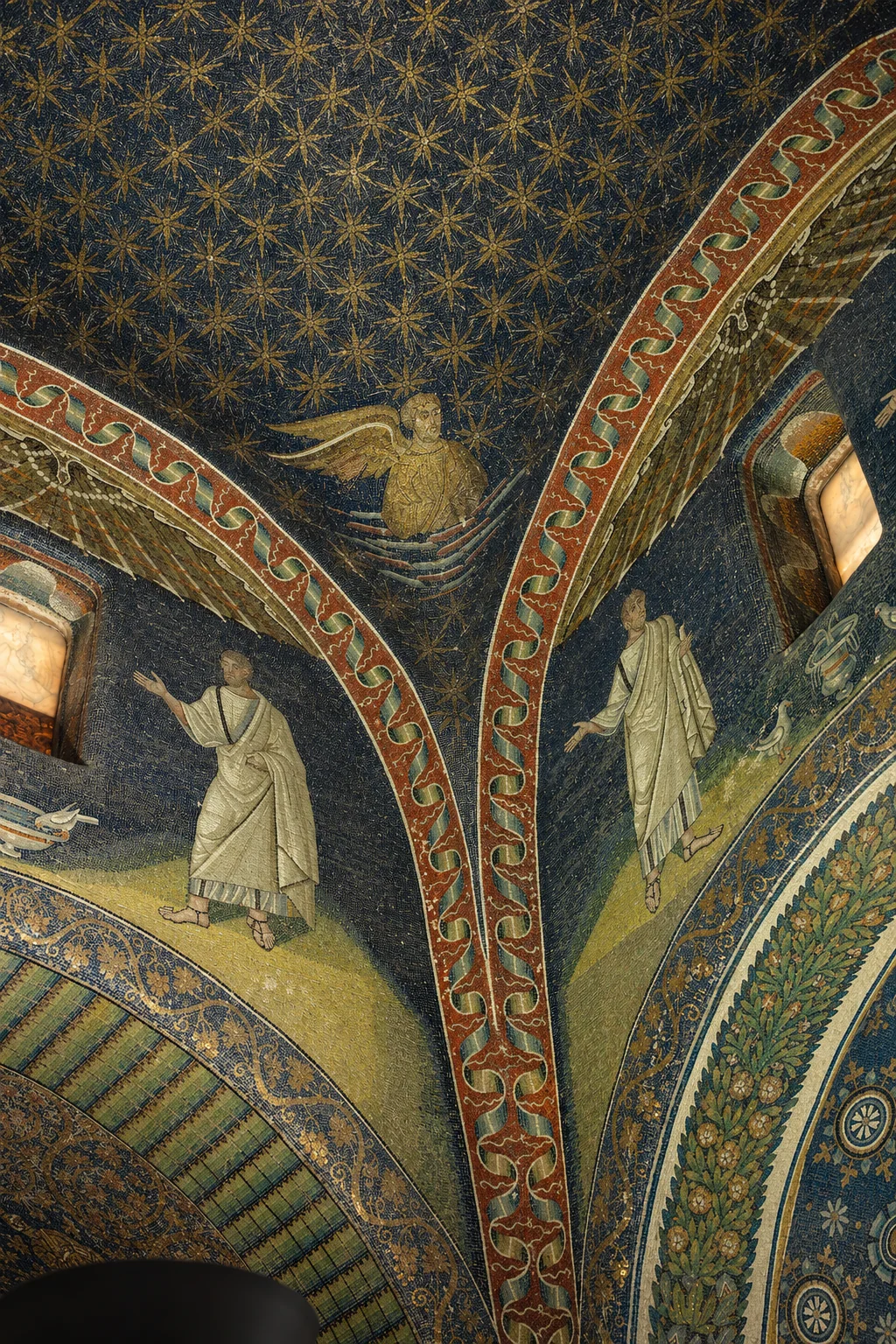 Gold stars on a blue dome above converging red ornamental arches, with pale-robed mosaic figures and partial alabaster windows below.