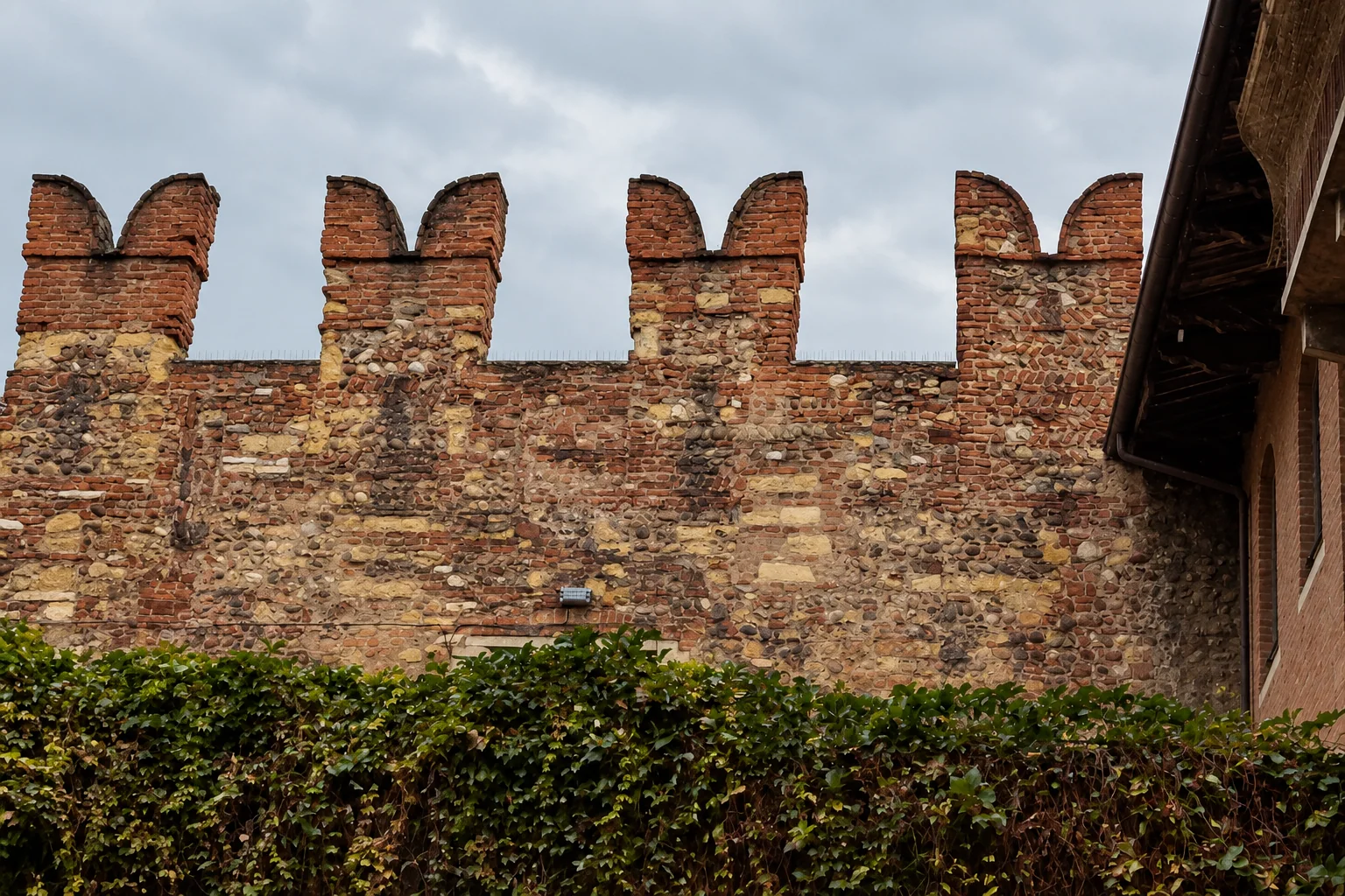 Four swallowtail brick merlons topping a rough stone courtyard wall, with ivy below and a roof edge at right.
