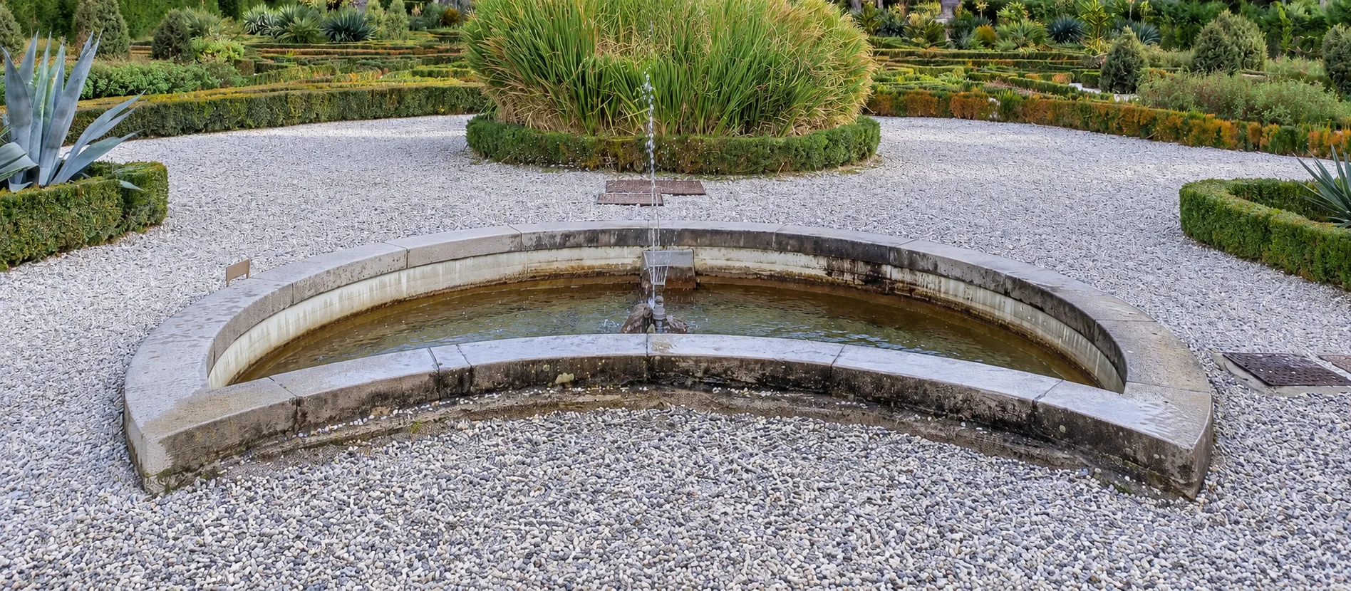Curved stone fountain basin with pale gravel, low clipped hedges and reeds in Miramare’s formal parterre.