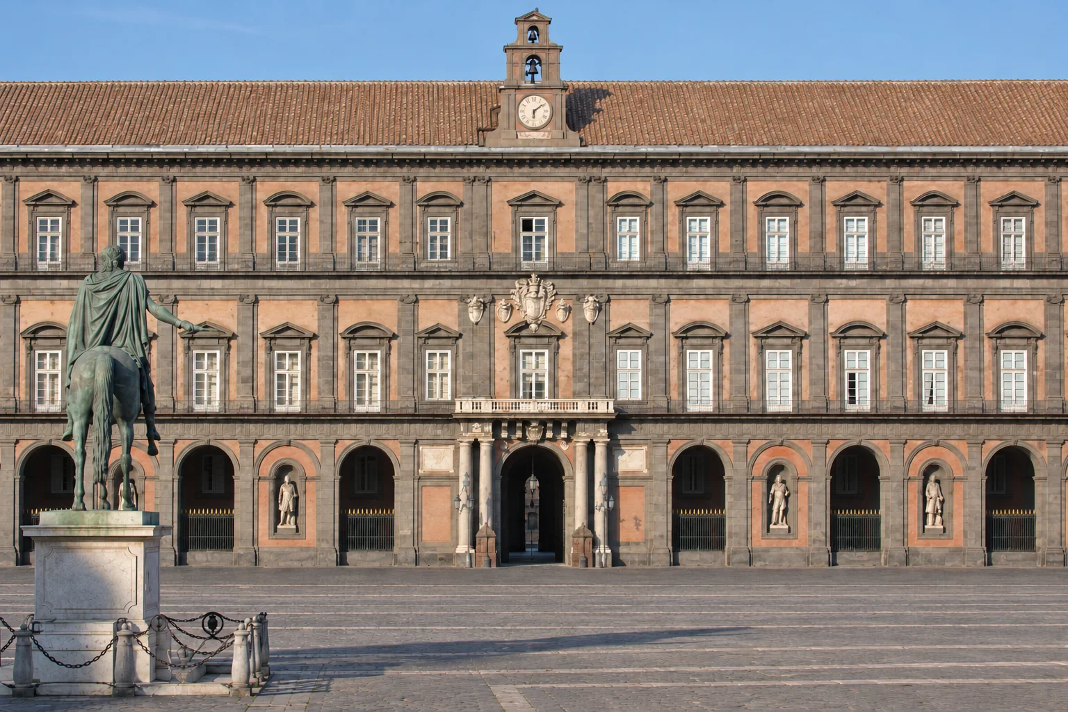 The Royal Palace’s long red-and-gray façade, with its repeated windows and ground-floor arcade, seen from Piazza del Plebiscito.