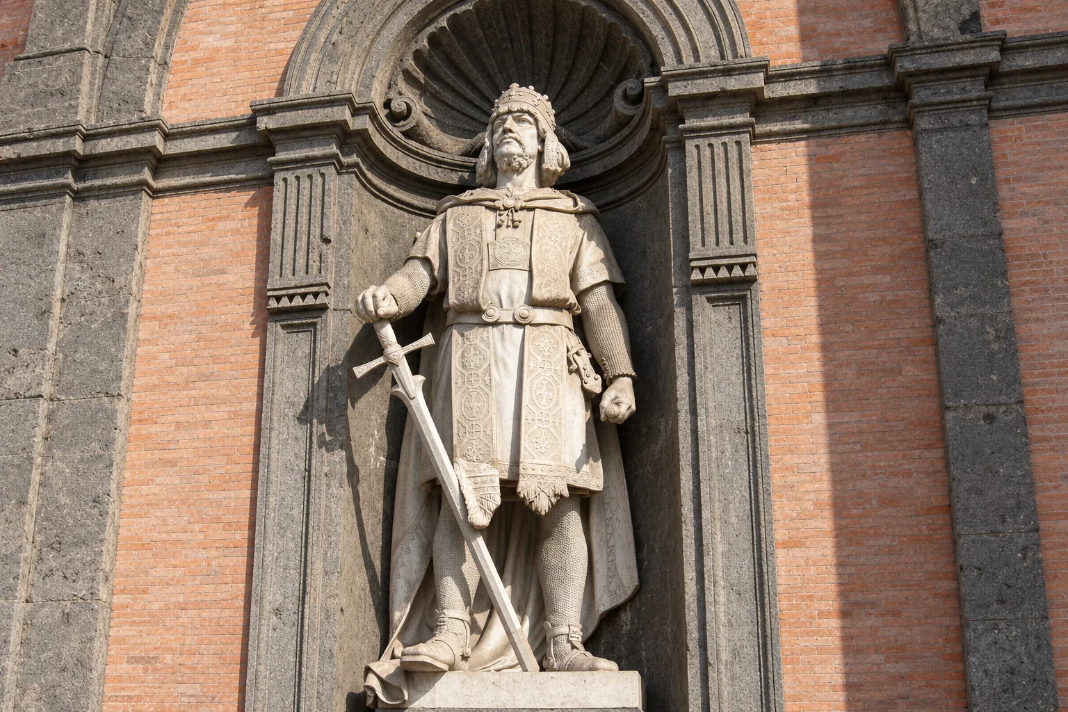 A crowned, bearded ruler statue holding a lowered sword inside a dark stone niche set in red brick.