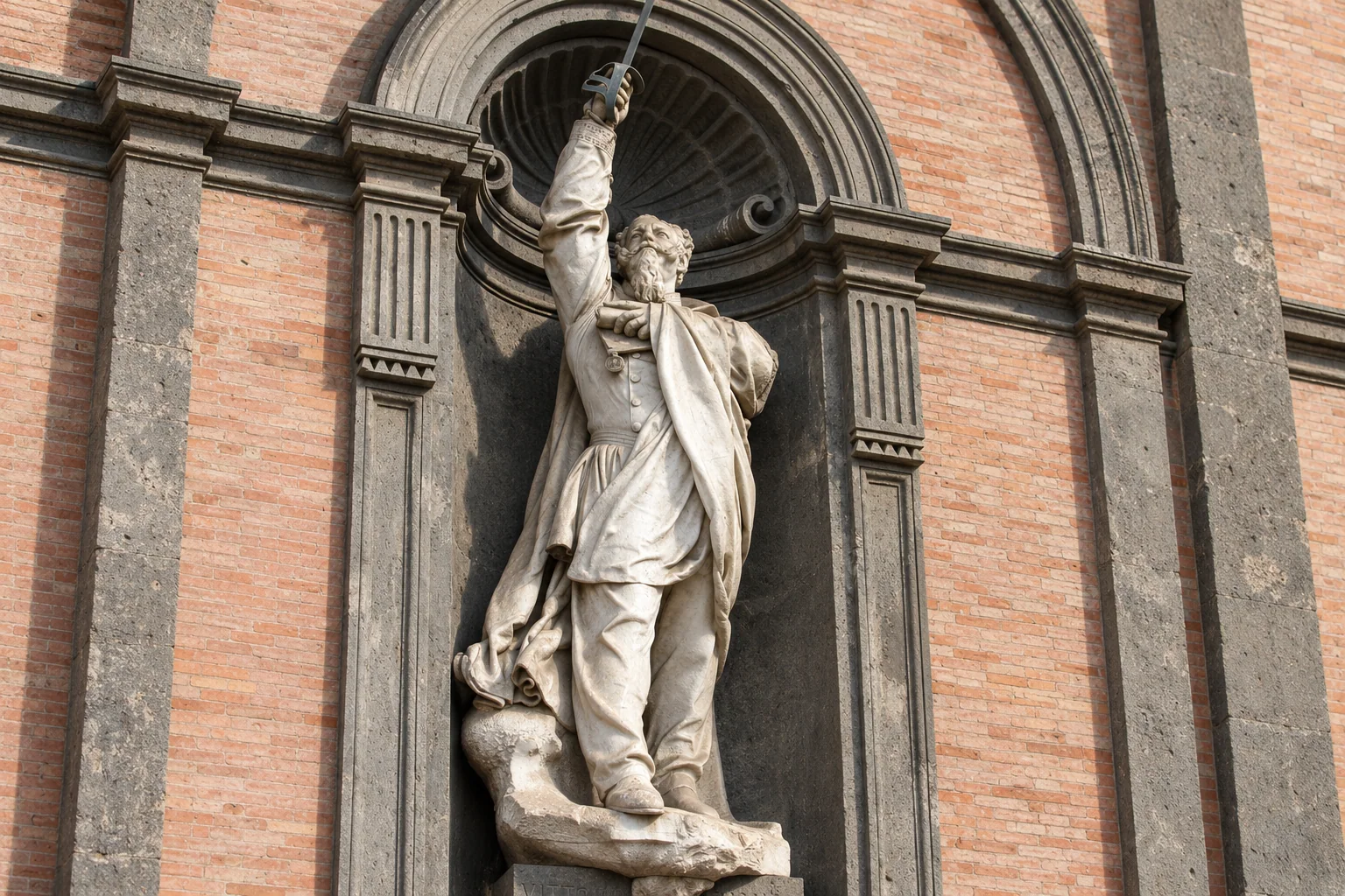 A full-length statue with one arm raised holding a sword, set in a rounded niche framed in dark stone and red brick.