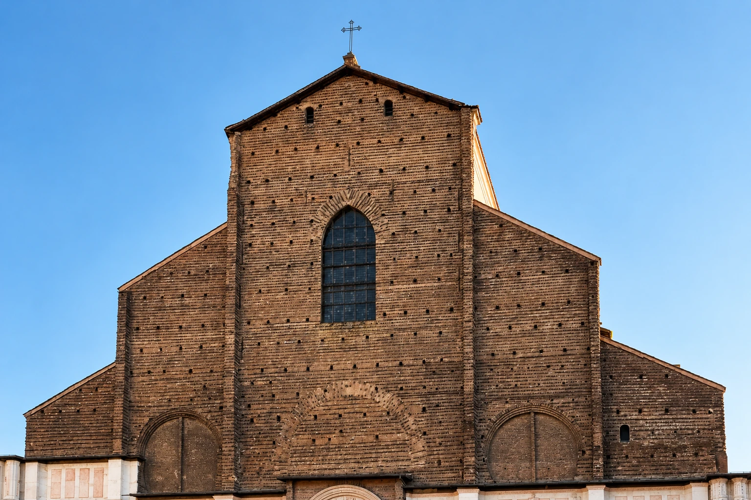 Upper brick façade of San Petronio, with a pointed central window, stepped roofline and slender iron cross.