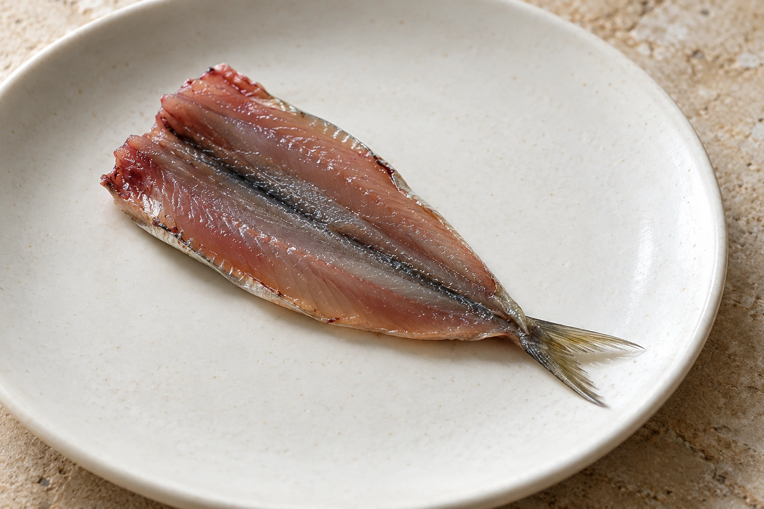 One headless raw sardine opened flat on a plate, with two connected flesh lobes and a single attached forked tail.