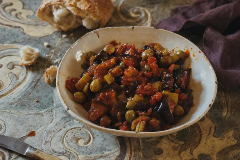 Caponata in a worn ceramic bowl on a faded majolica table, glossy eggplant, celery and green olives in dark sweet and sour glaze, torn bread above.