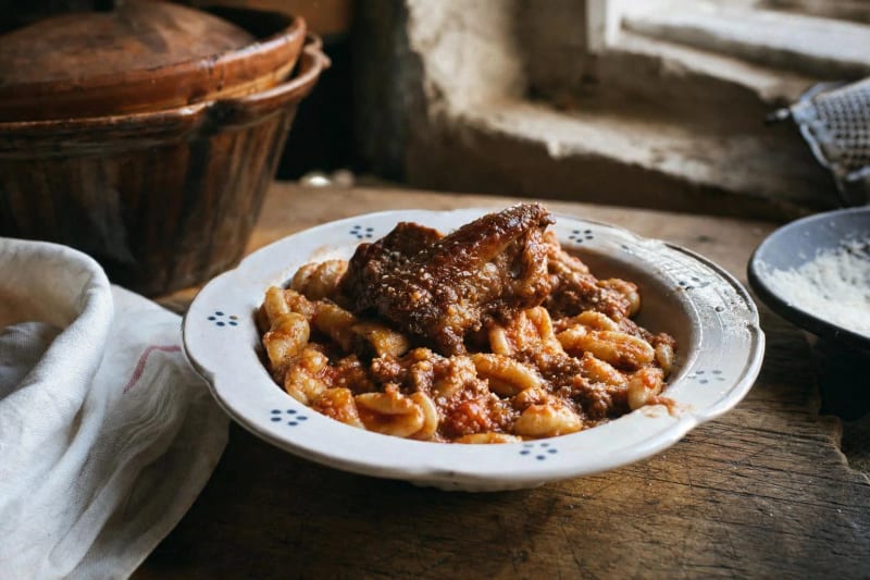 Cavatelli al ragu di maiale in a dotted bowl, hand-curled shells in brick red sauce under a glossy braised rib, pecorino at the window.