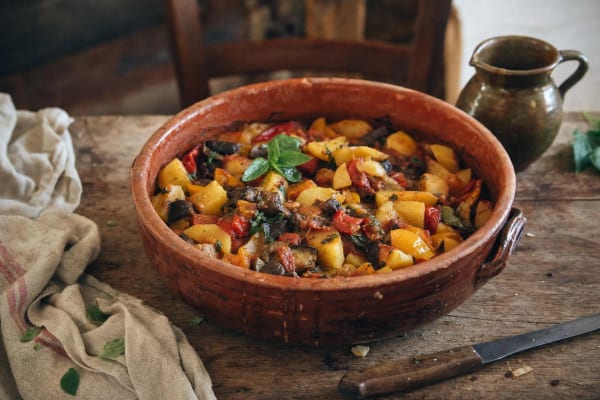 Ciambotta lucana in a wide terracotta dish, golden potatoes, red peppers and dark eggplant still in distinct pieces under torn basil, green jug behind.