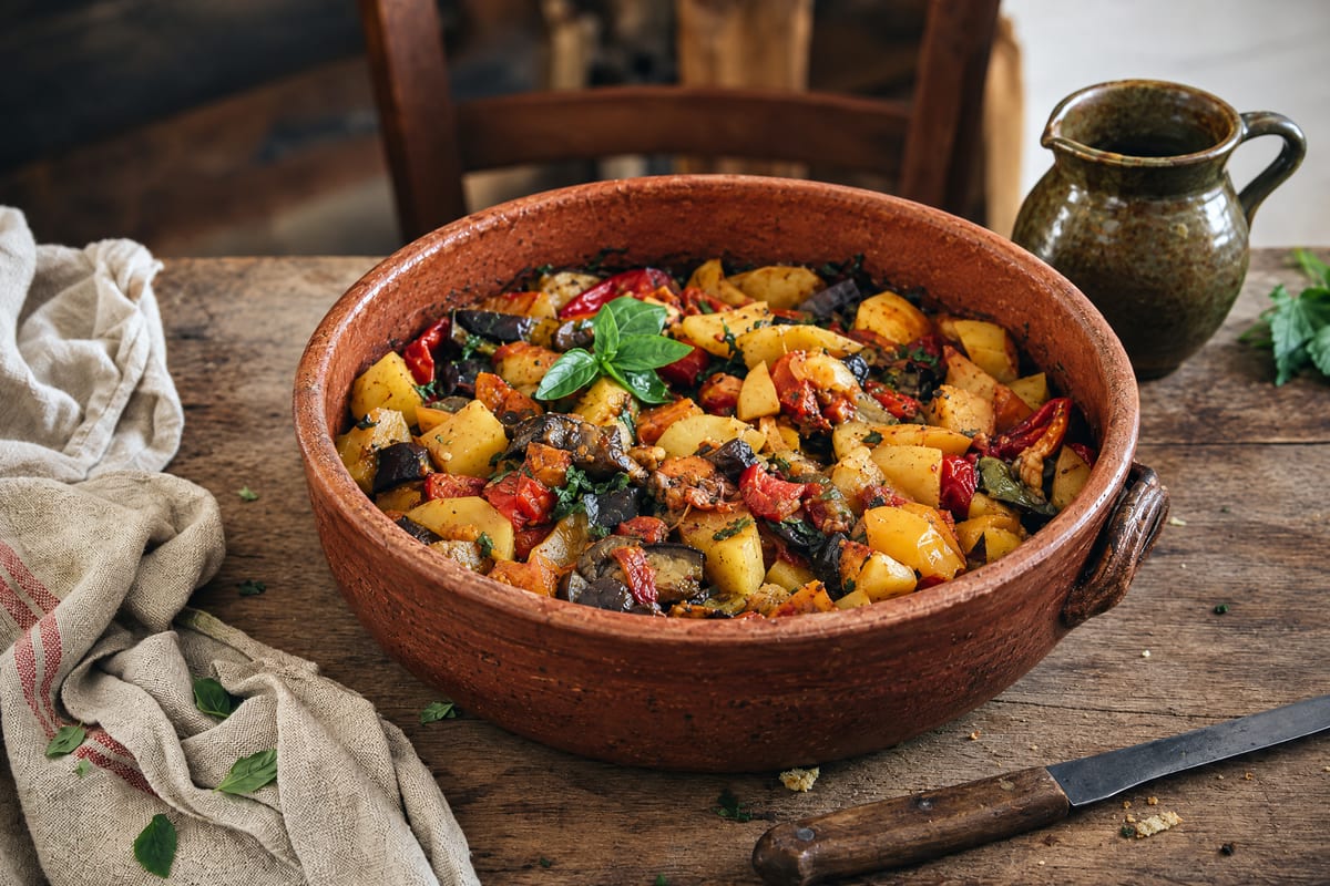 Ciambotta lucana in a wide terracotta dish, golden potatoes, red peppers and dark eggplant still in distinct pieces under torn basil, green jug behind.