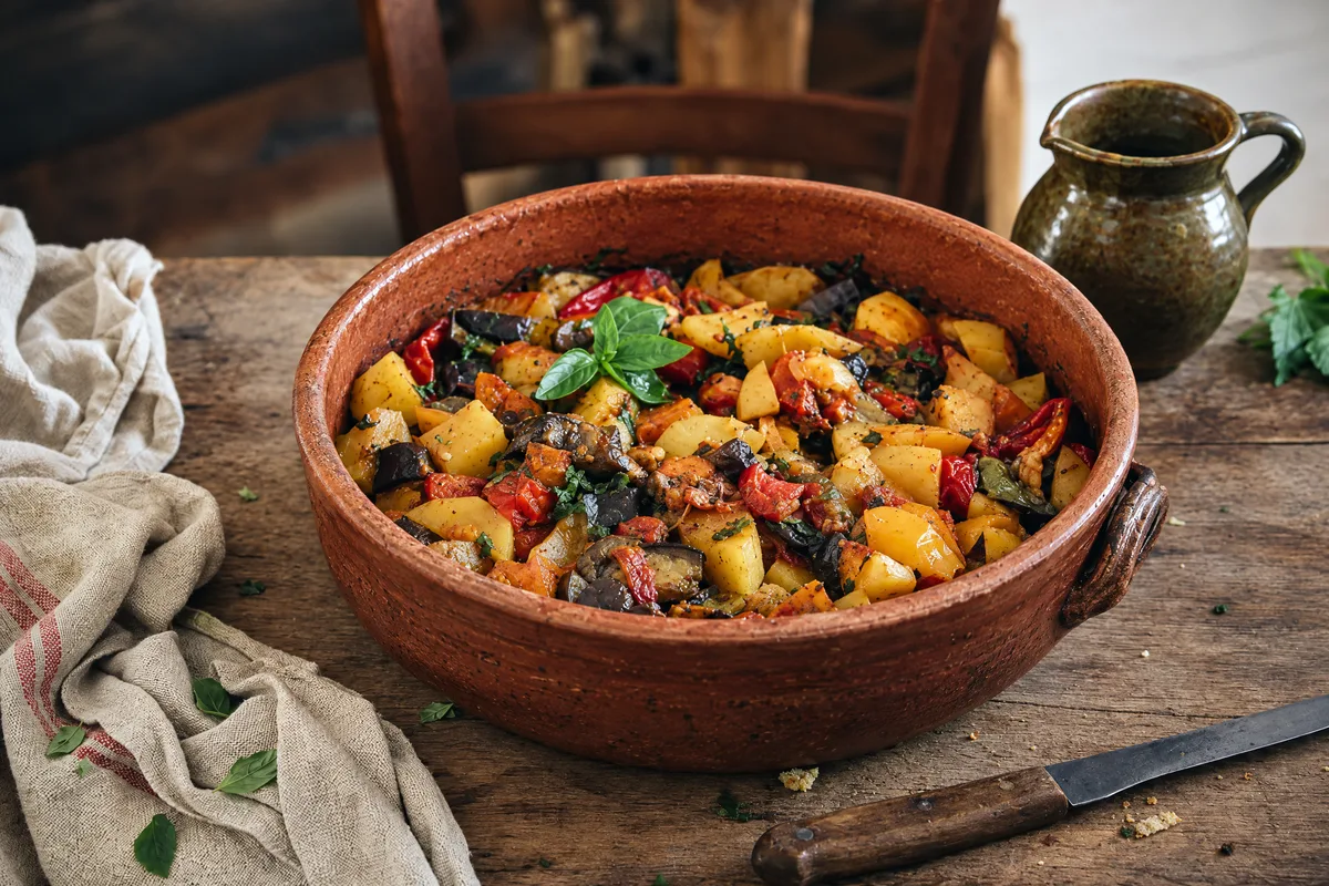 Ciambotta lucana in a wide terracotta dish, golden potatoes, red peppers and dark eggplant still in distinct pieces under torn basil, green jug behind.
