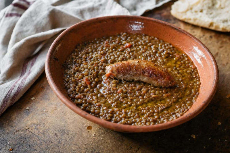 Lenticchie di Castelluccio in umido in a terracotta bowl, glossy tiny lentils with a ring of olive oil and a browned sausage resting on top.