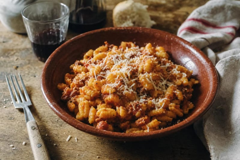 Malloreddus alla campidanese in a terracotta bowl, ridged saffron gnocchetti in sausage ragu under a drift of pecorino, red wine and a fork beside.