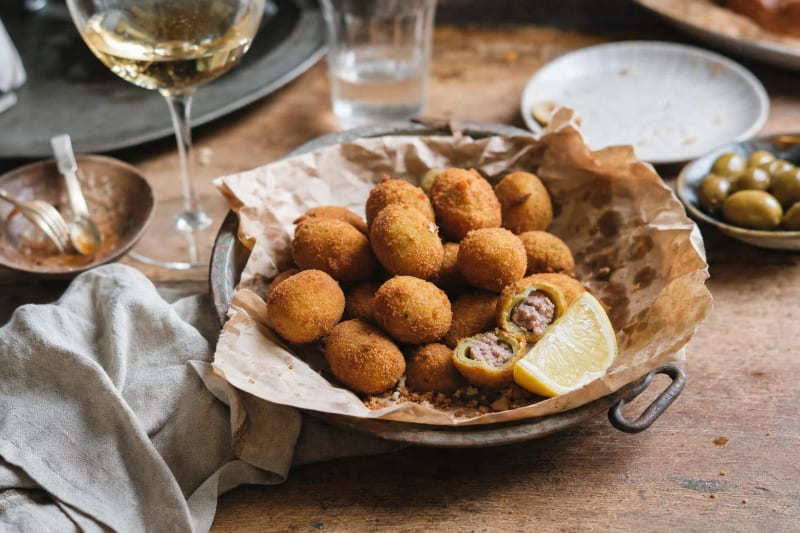 Olive all ascolana heaped on paper in a tin dish, golden fried stuffed olives with one broken open to its meat filling, lemon wedge and white wine near.