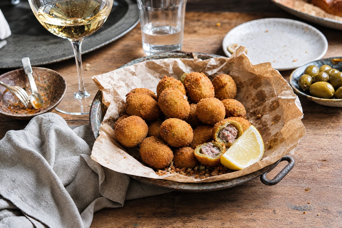 Olive all ascolana heaped on paper in a tin dish, golden fried stuffed olives with one broken open to its meat filling, lemon wedge and white wine near.