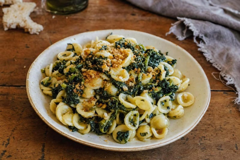 Orecchiette alle cime di rapa on a cream plate, pasta ears tangled with dark turnip tops under golden toasted breadcrumbs, oil and bread above.
