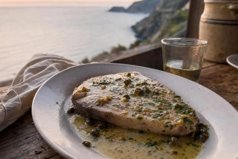 Pesce spada alla bagnarese, a pale golden pan-cooked swordfish steak under salmoriglio with capers and parsley, white wine and the Costa Viola at dusk behind.