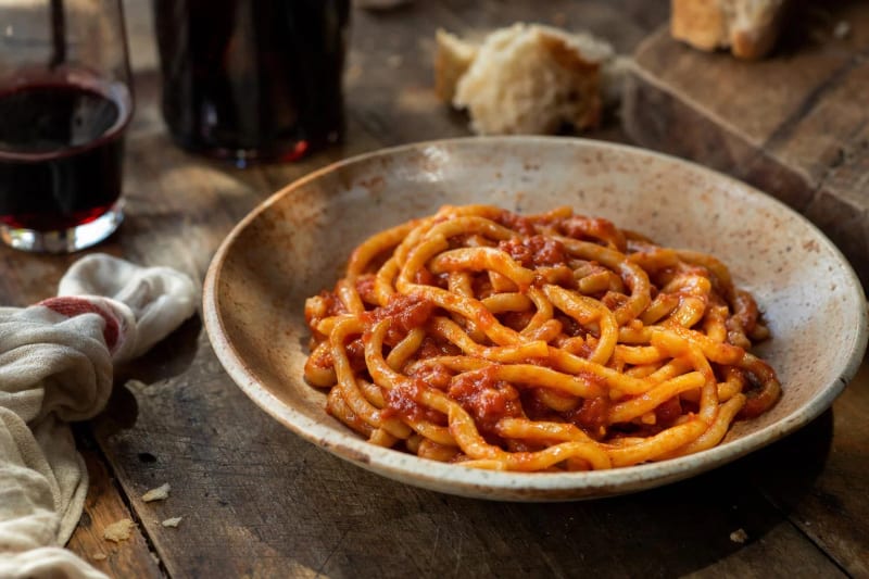 Pici all aglione coiled in a speckled bowl, the thick hand-rolled noodles gloved in deep red garlic and tomato sauce, bread and red wine behind.