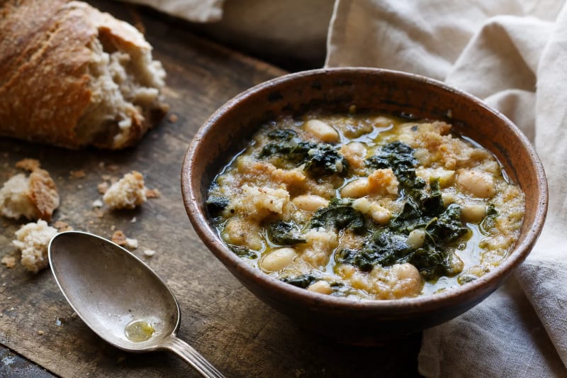 Ribollita in a rustic ceramic bowl, thick with cavolo nero, cannellini and torn bread under a ring of olive oil, crumbs and a worn spoon beside.