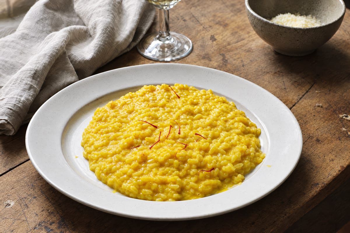 Risotto alla milanese spread loose on a white plate, saffron gold with a few crimson threads on top, grated cheese and a glass of wine behind.