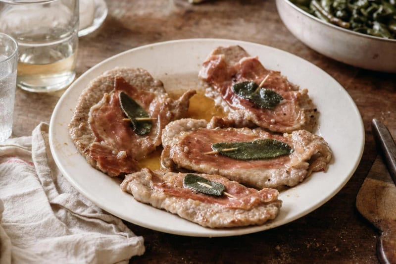 Saltimbocca alla romana on a white plate, flat veal cutlets each pinned with prosciutto and a sage leaf in a thin pan glaze, green beans behind.