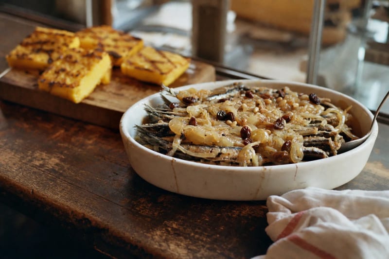 Sarde in saor in an oval dish, fried sardines buried under soft golden onions with raisins and pine nuts, squares of grilled polenta on a board behind.