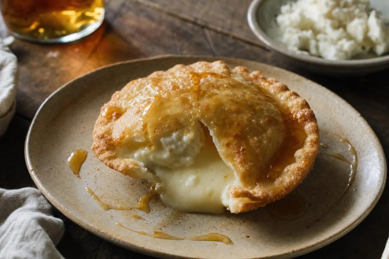 A seada on a stoneware plate, the fried pastry round broken open with young pecorino flowing out under ribbons of amber honey, mirto glowing behind.
