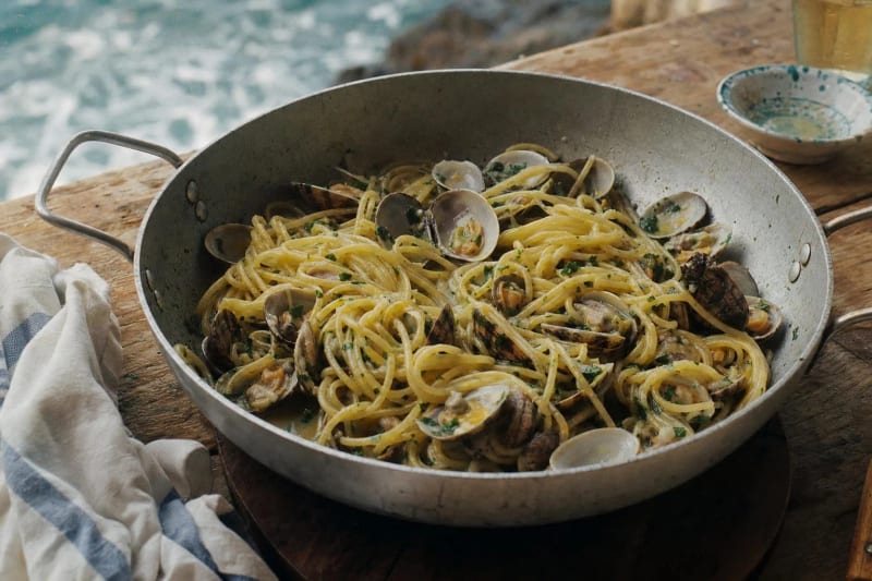 Spaghetti alle vongole in bianco in a battered aluminum pan, open carpet-shell clams through glossy strands flecked with parsley, the sea breaking below.