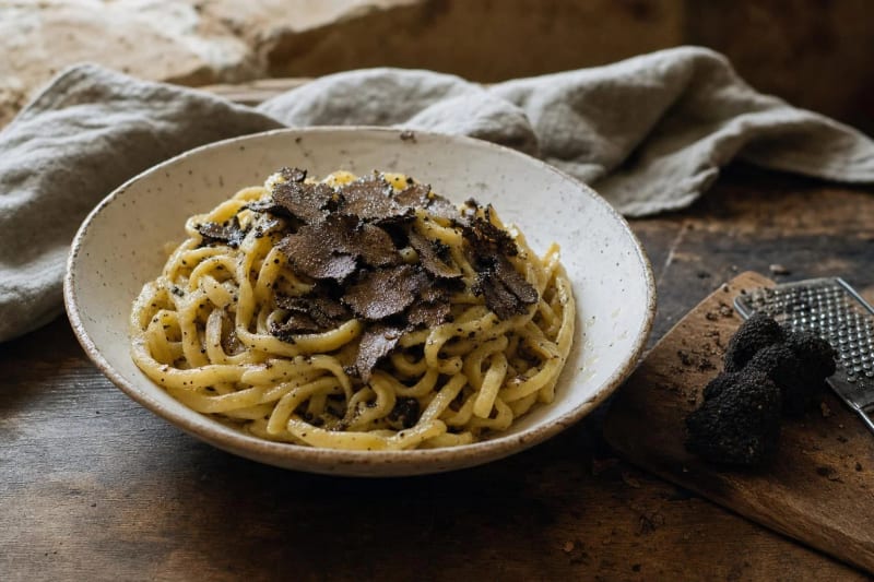 Strangozzi al tartufo nero, pasta ribbons flecked with truffle under a crown of dark shavings, whole black truffles and a grater on the board.