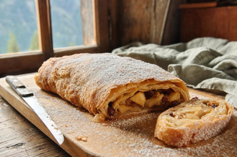 Strudel di mele on a floured board by an alpine window, icing sugar over the blistered pastry and a cut slice showing the spiral of apples and raisins.