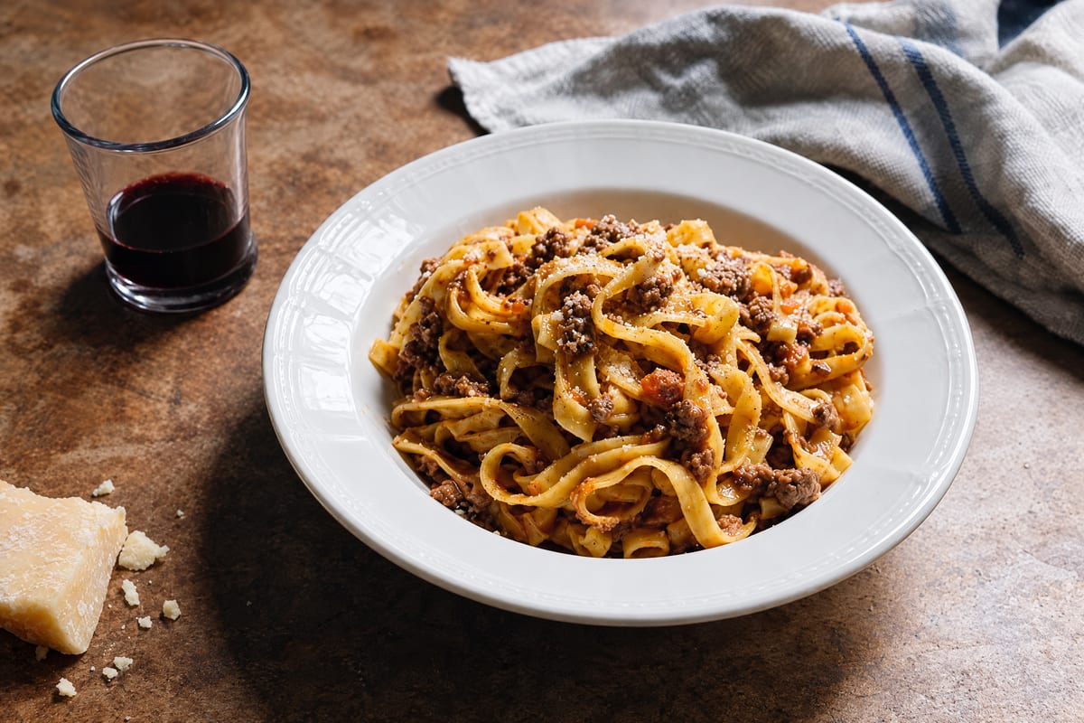 Tagliatelle al ragu bolognese piled in a cream bowl, the slow meat sauce clinging to golden ribbons with barely a blush of tomato, Parmigiano and red wine near.