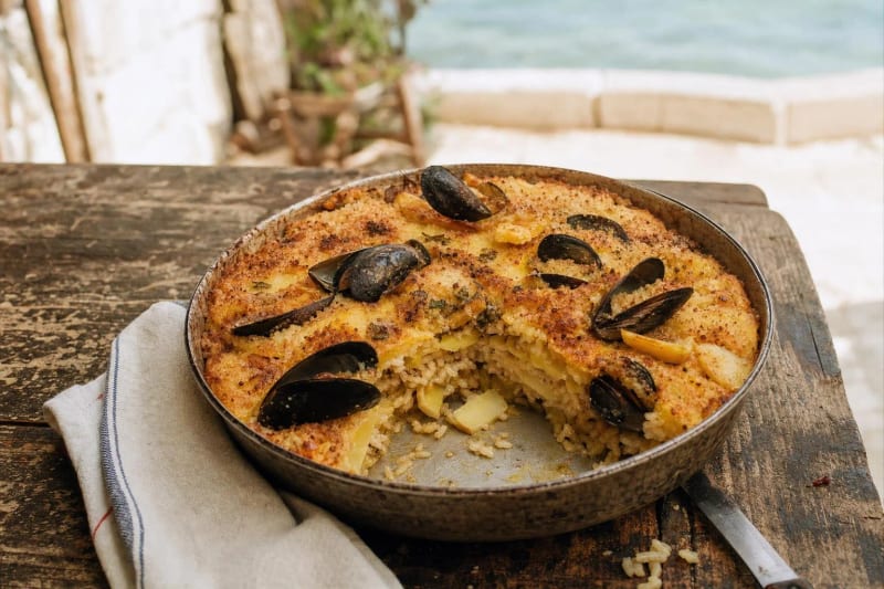 Tiella barese in its round pan with a wedge lifted out, bronzed crust over rice, potatoes and mussels on their half shells, Bari seawater beyond.