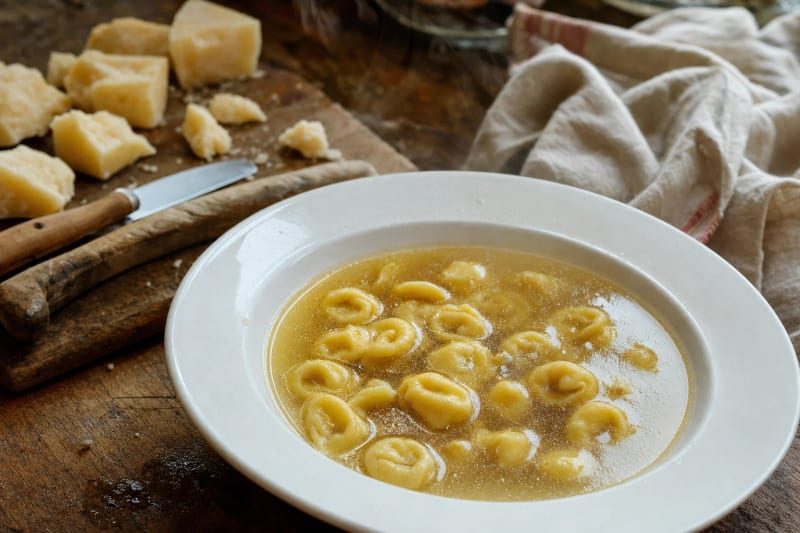 Tortellini in brodo in a white rimmed bowl, small hand-folded pasta floating in clear golden capon broth, chunks of Parmigiano on the board behind.