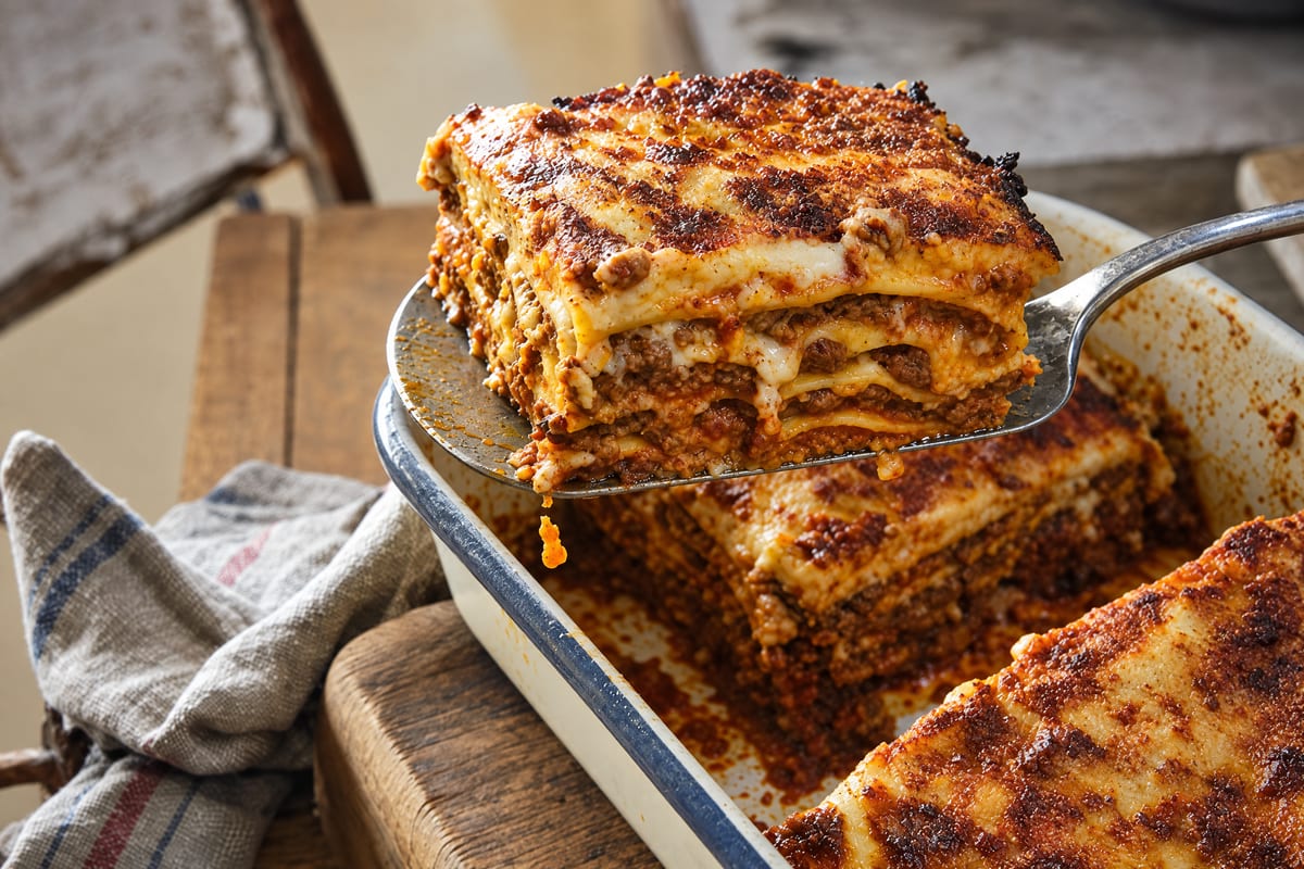 Vincisgrassi lifted from an enamel baking dish on a worn spatula, the burnished square showing dark ragu and besciamella layers, corner crust trailing.